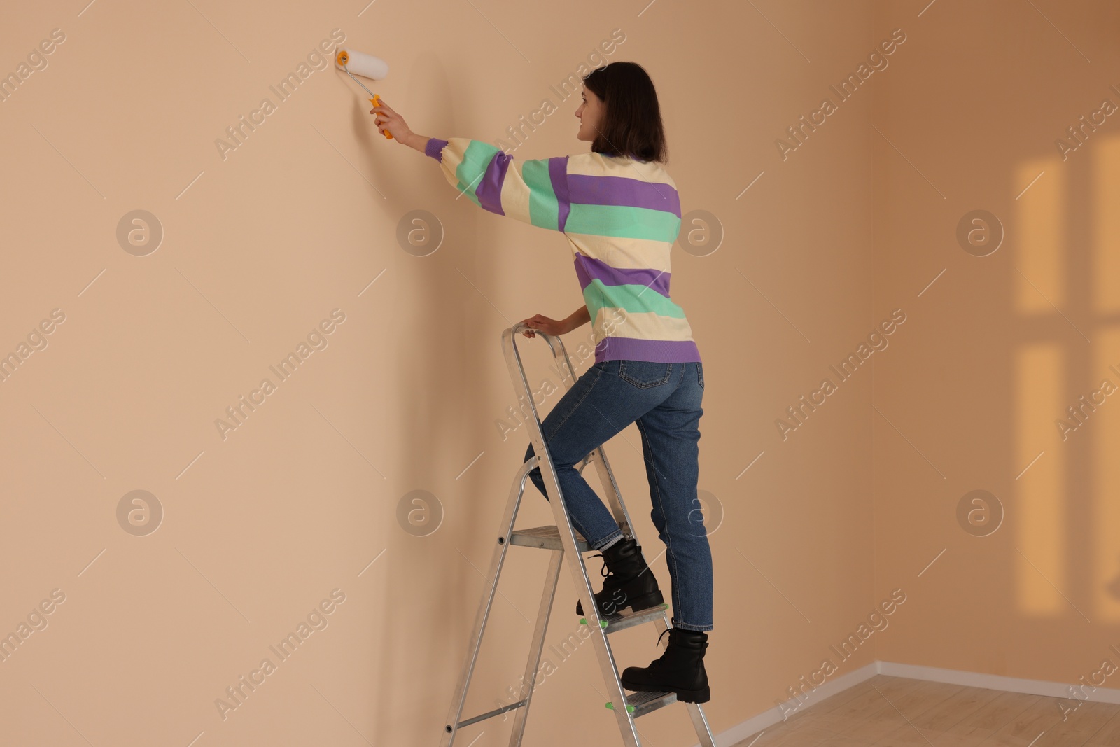 Photo of Young woman painting wall with roller on stepladder indoors. Room renovation