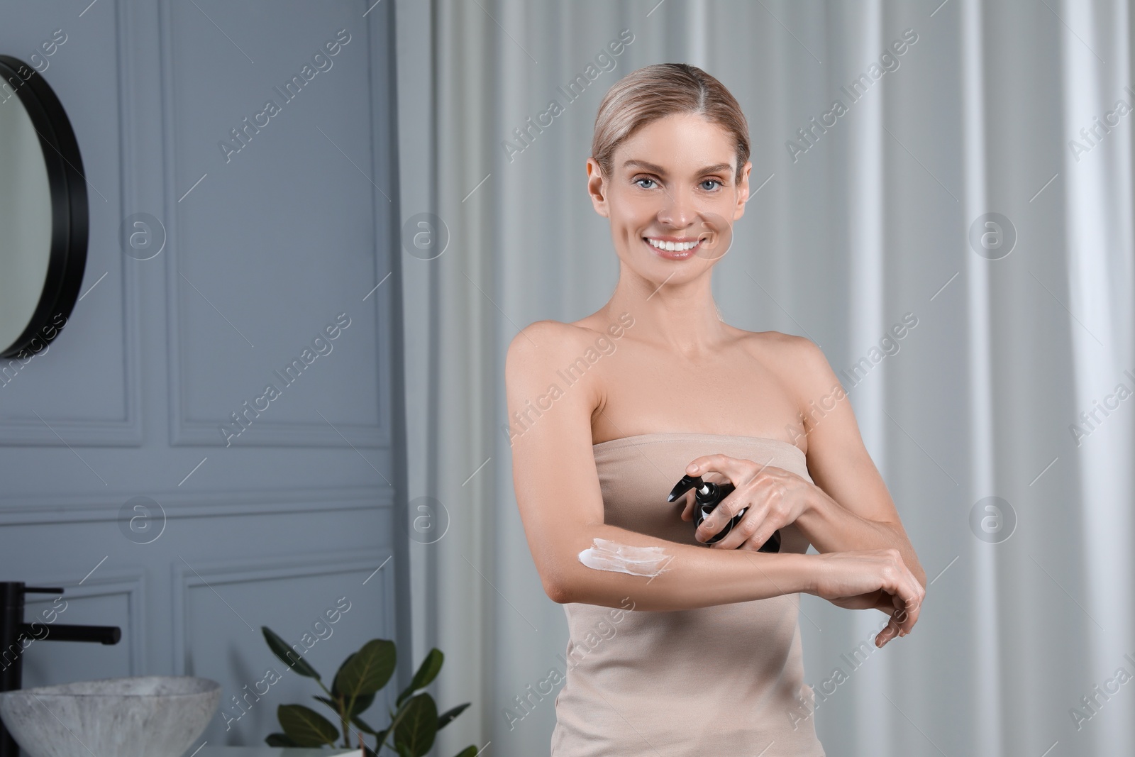 Photo of Happy woman applying body cream onto arm in bathroom, space for text