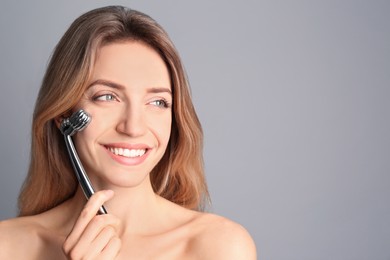 Young woman using metal face roller on light grey background, space for text