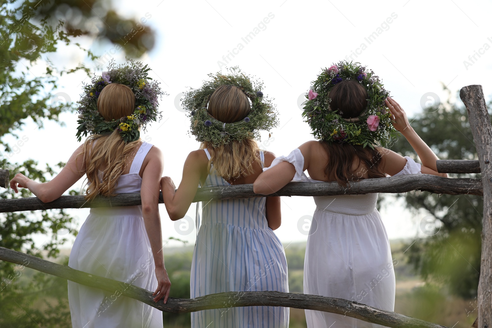 Photo of Young women wearing wreaths made of beautiful flowers near wooden fence, back view