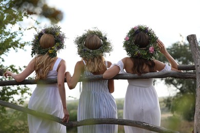 Young women wearing wreaths made of beautiful flowers near wooden fence, back view