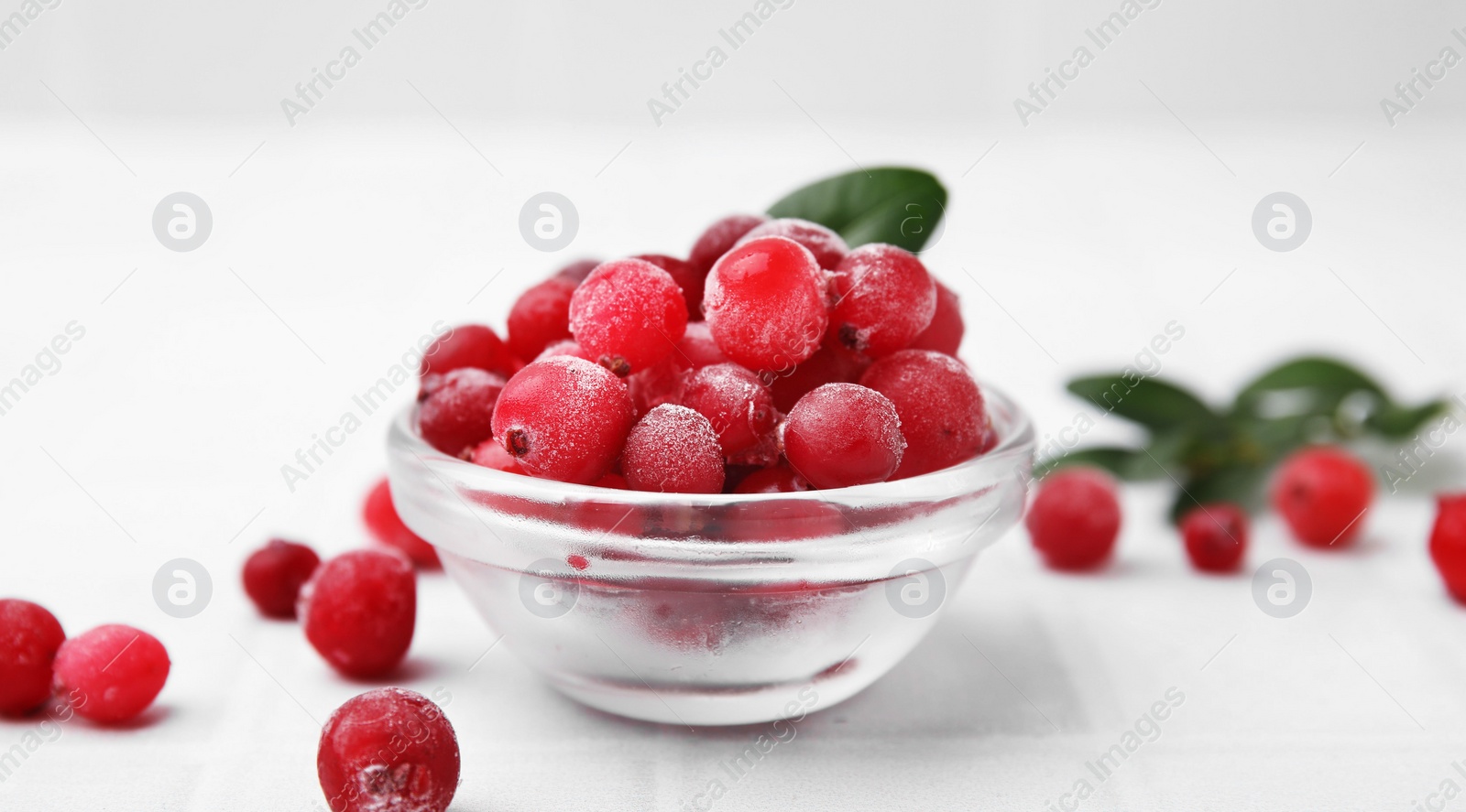 Photo of Frozen red cranberries in bowl and green leaves on white table, closeup