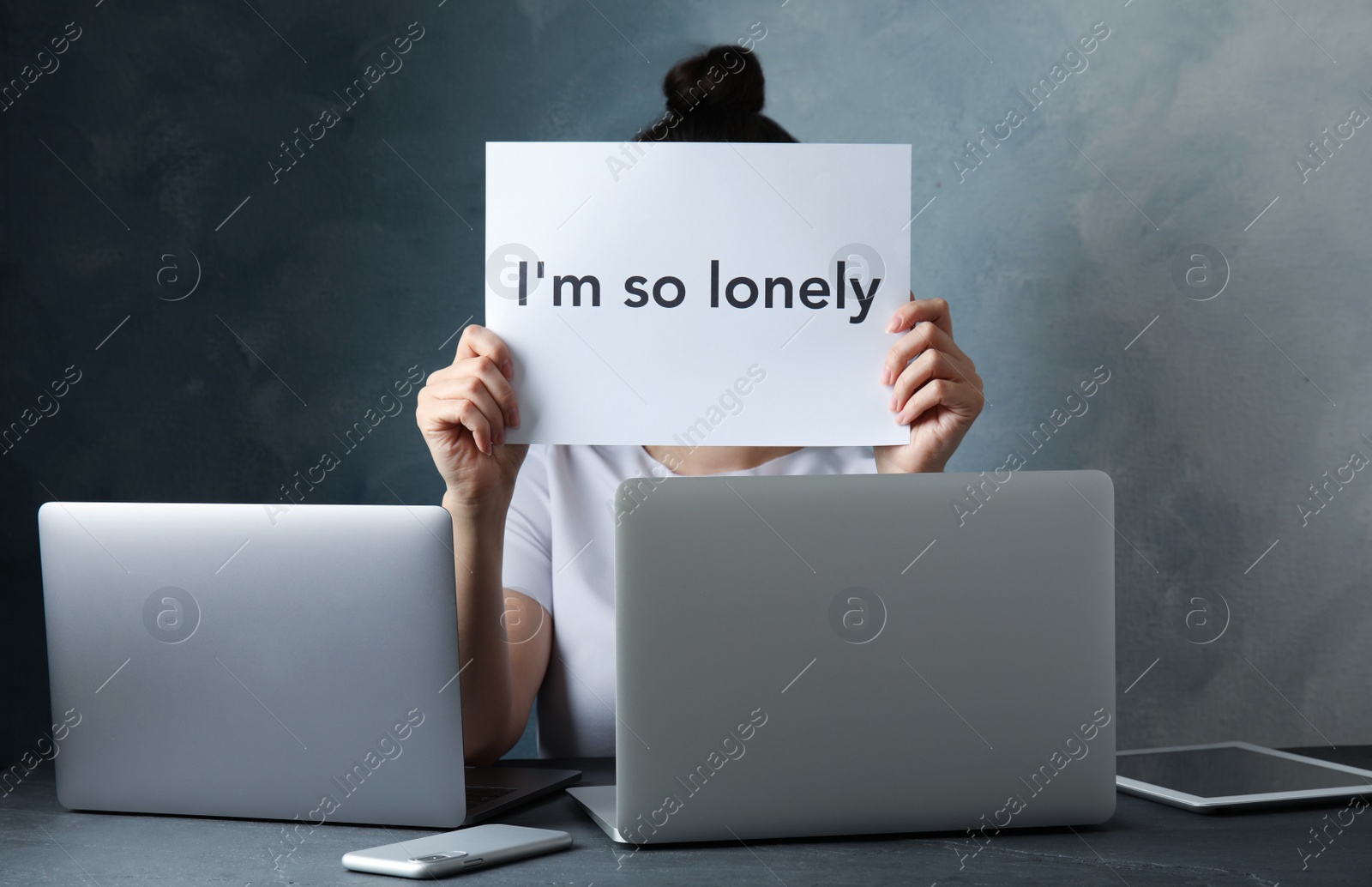 Photo of Woman with note I'M SO LONELY and gadgets at table against black background. Solitude concept