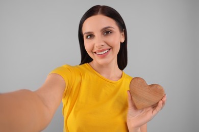Photo of Happy young woman with decorative wooden heart taking selfie on grey background