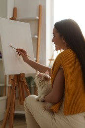Photo of Beautiful young woman drawing on easel with cat at home
