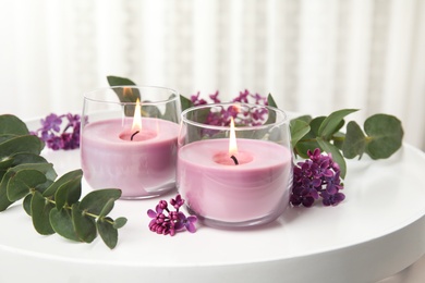 Burning candles in glass holders and flowers with leaves on white table