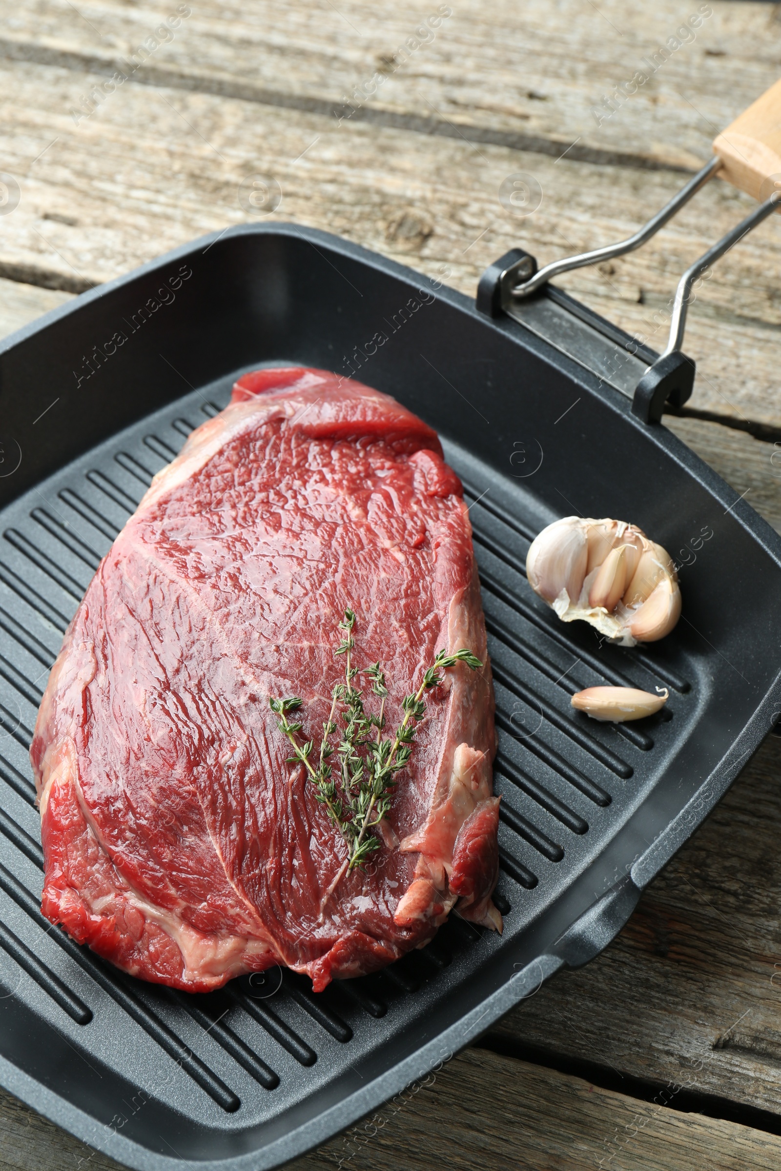 Photo of Grill pan with piece of raw beef meat, garlic and thyme on wooden table