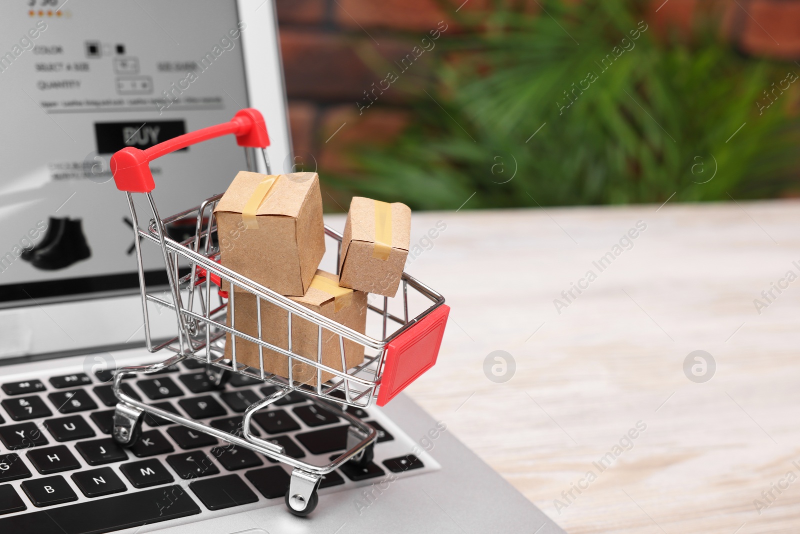 Photo of Internet store. Small cardboard boxes, shopping cart and laptop on light wooden table, closeup. Space for text