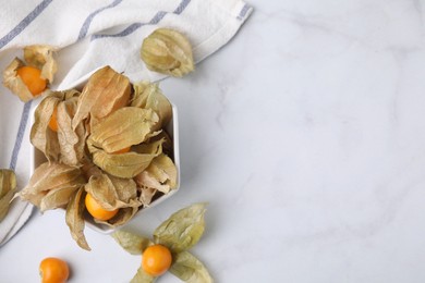 Ripe physalis fruits with calyxes in bowl on white marble table, flat lay. Space for text