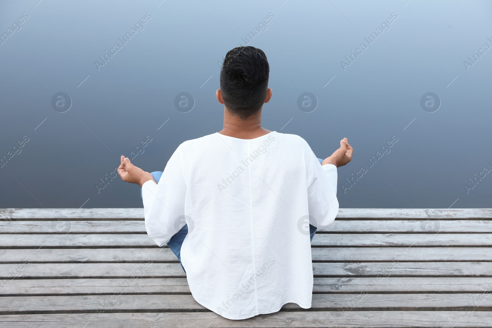 Photo of Man meditating on wooden pier near river, back view