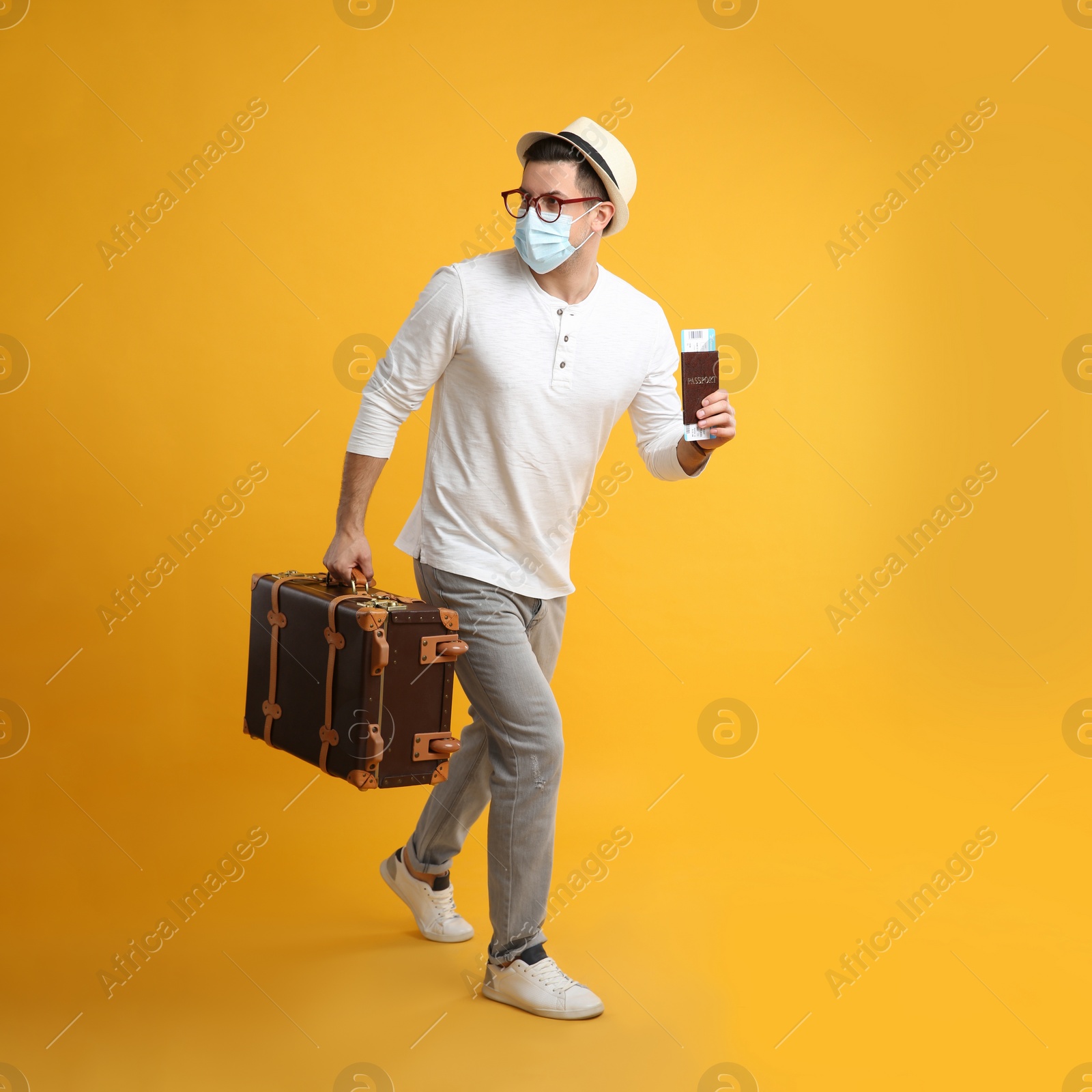 Photo of Male tourist n protective mask holding passport with ticket and suitcase on yellow background
