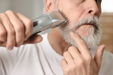 Photo of Man trimming beard with electric trimmer indoors, closeup