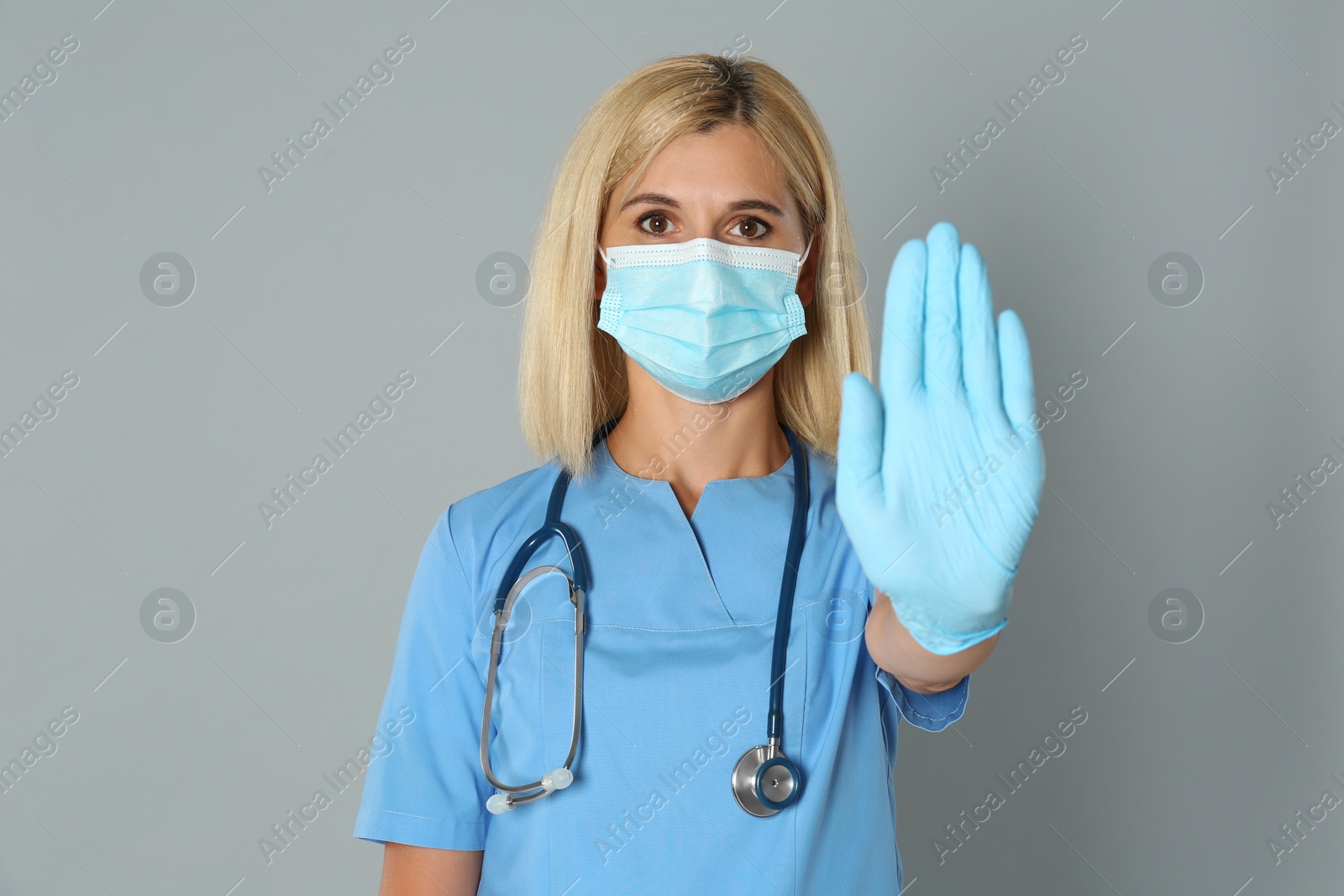 Photo of Doctor in protective mask showing stop gesture on grey background. Prevent spreading of coronavirus