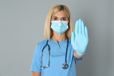 Photo of Doctor in protective mask showing stop gesture on grey background. Prevent spreading of coronavirus