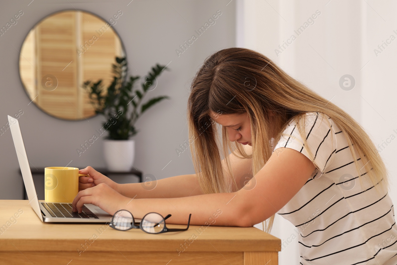 Photo of Sleepy young woman with cup of coffee and laptop at wooden table indoors