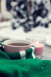 Photo of Winter morning. Cup with hot drink wrapped in green sweater and burning candle outdoors, closeup. Space for text
