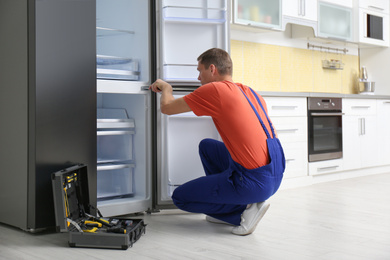 Male technician with screwdriver repairing refrigerator in kitchen