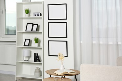 Photo of Stylish room interior with empty frames hanging on white wall near shelving unit