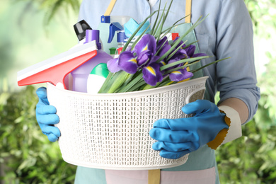 Photo of Woman holding basket with spring flowers and cleaning supplies on green blurred background, closeup