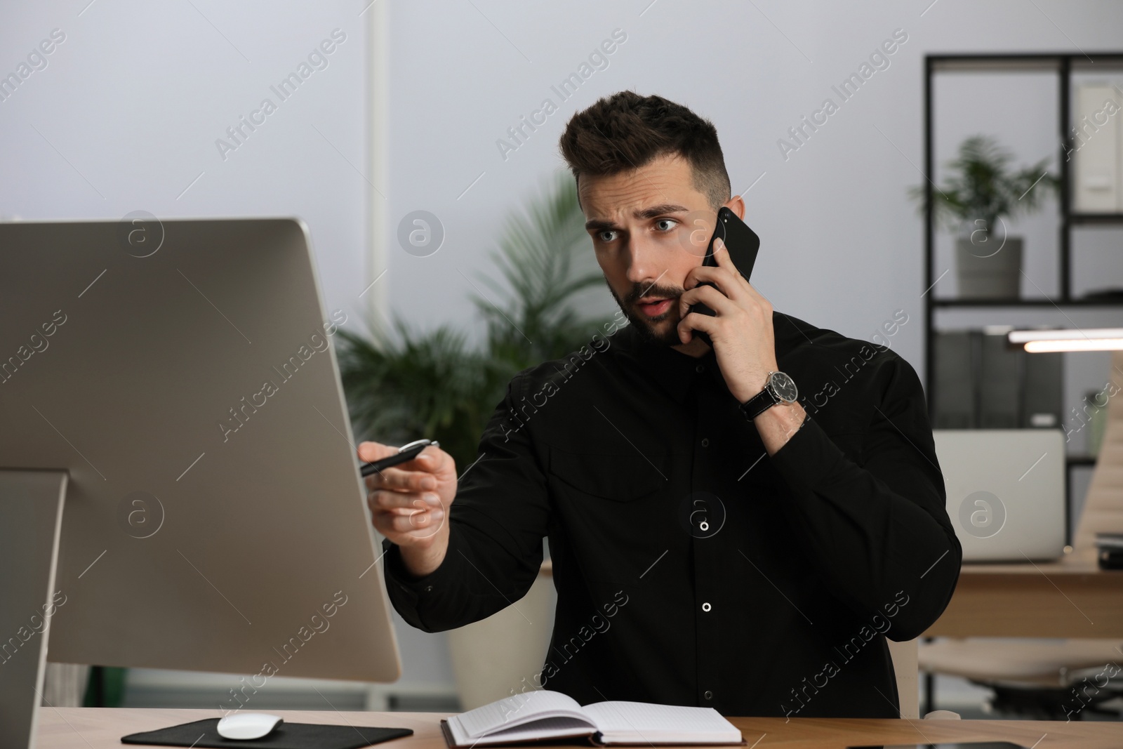 Photo of Man talking on phone while working with computer at table in office