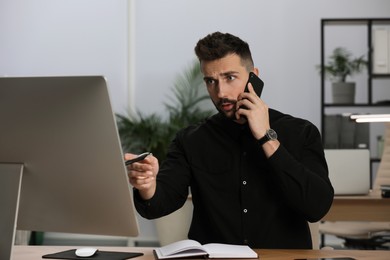 Photo of Man talking on phone while working with computer at table in office