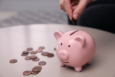 Photo of Woman putting coin into piggy bank at table indoors, closeup