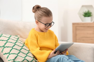 Cute little girl in glasses with tablet on sofa indoors