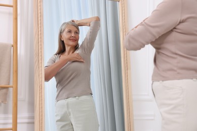 Photo of Beautiful senior woman doing breast self-examination near mirror indoors