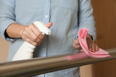 Photo of Woman cleaning metal railing with rag and spray detergent indoors, closeup