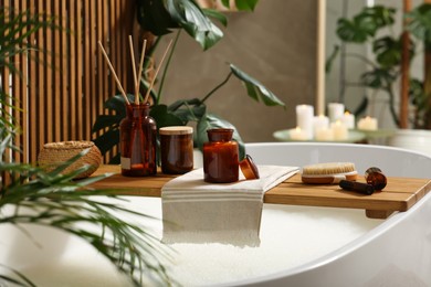 Photo of Wooden bath tray with open book, candle and body care products on tub indoors. Relaxing atmosphere