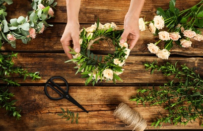 Photo of Top view of woman making wreath with beautiful flowers at wooden table, closeup