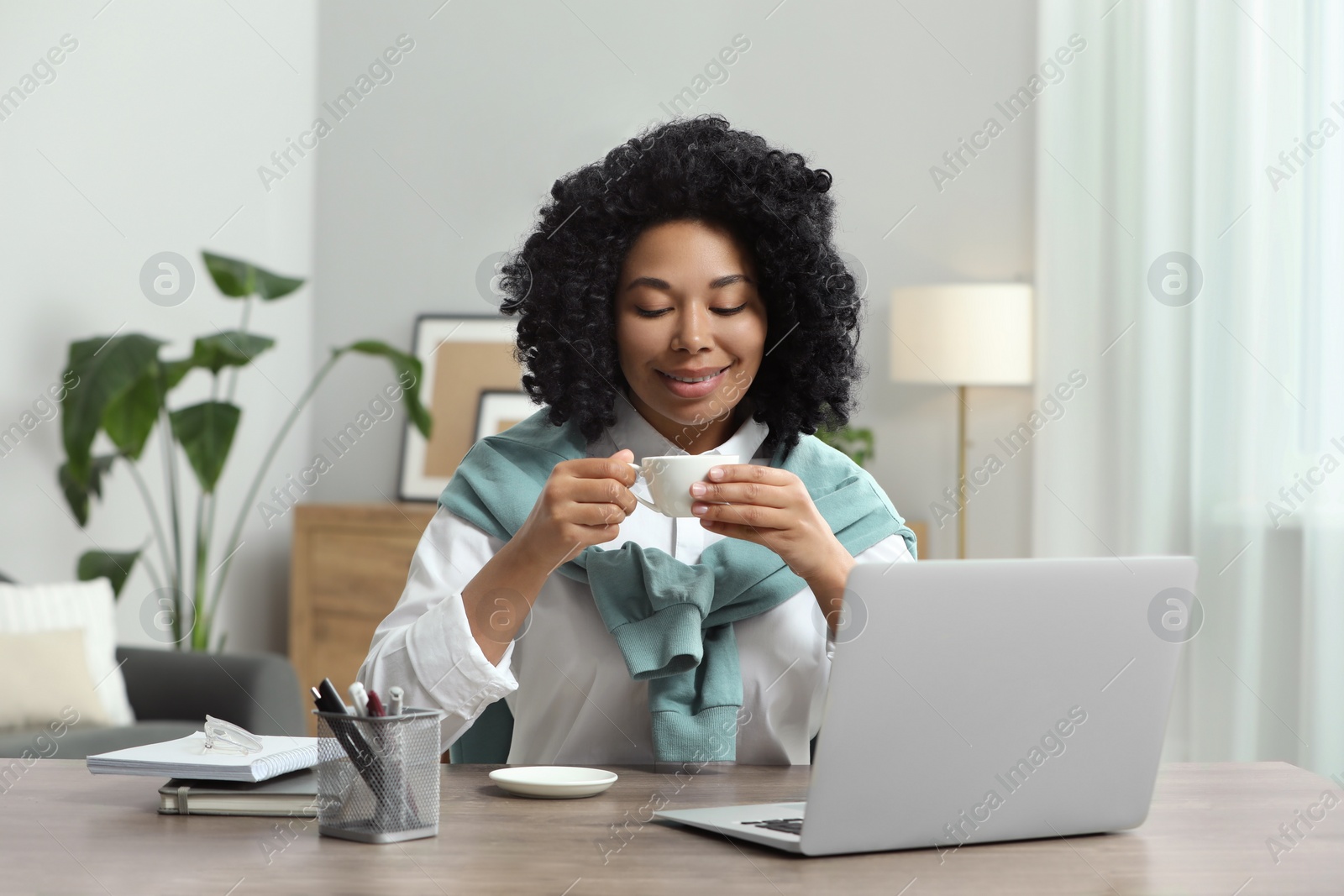 Photo of Happy young woman with cup of drink using laptop at wooden desk indoors