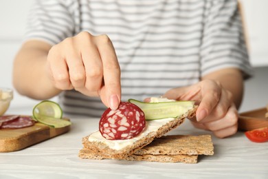 Photo of Woman making sandwich with rye crispbread, cream cheese, sausage and cucumber slice at white wooden table, closeup