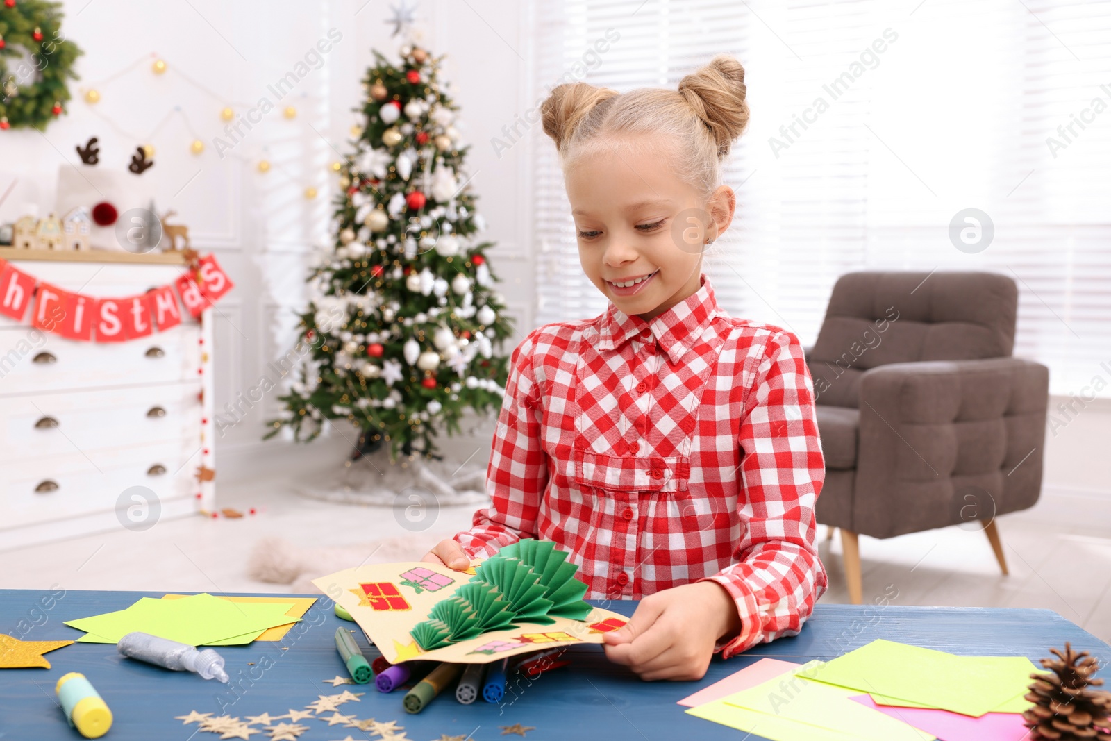 Photo of Cute little child with beautiful Christmas card at table in decorated room