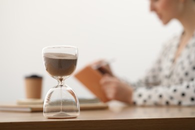 Photo of Hourglass with flowing sand on desk. Woman working indoors, selective focus