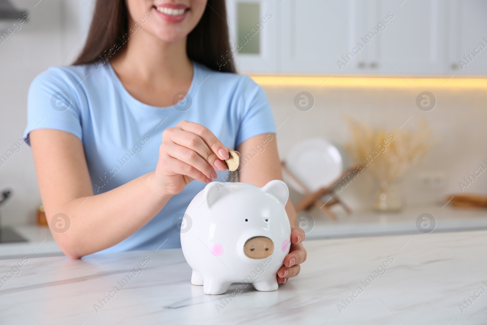 Photo of Young woman putting coin into piggy bank at table indoors, closeup. Money savings