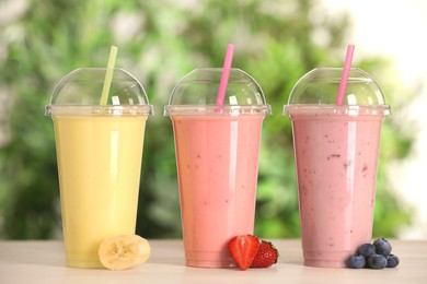 Photo of Plastic cups with different tasty smoothies and fresh fruits on wooden table outdoors