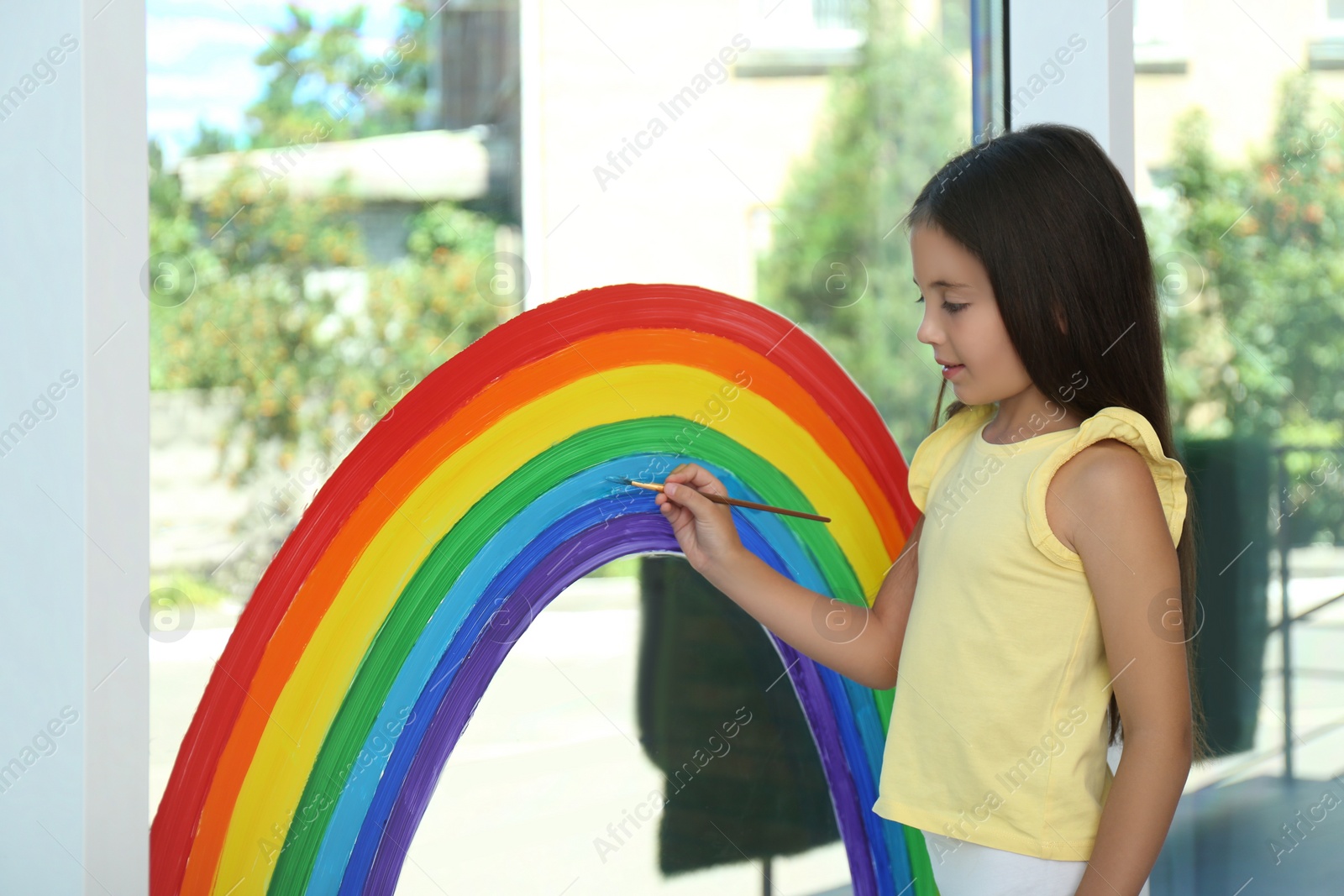 Photo of Little girl drawing rainbow on window indoors. Stay at home concept