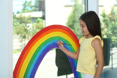 Little girl drawing rainbow on window indoors. Stay at home concept