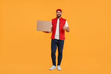 Photo of Happy young courier with parcel showing thumb up on orange background