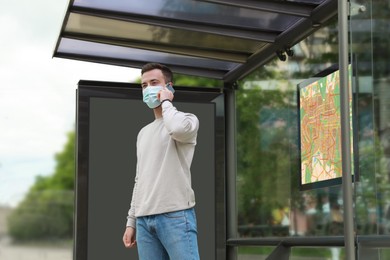 Image of Young man in protective mask talking on phone while waiting for public transport at bus stop