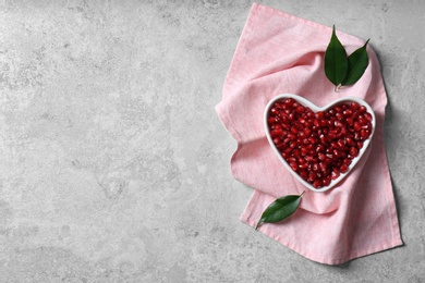 Heart shaped bowl of pomegranate seeds on grey background, top view with space for text