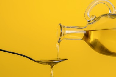 Pouring cooking oil from jug into spoon on yellow background, closeup
