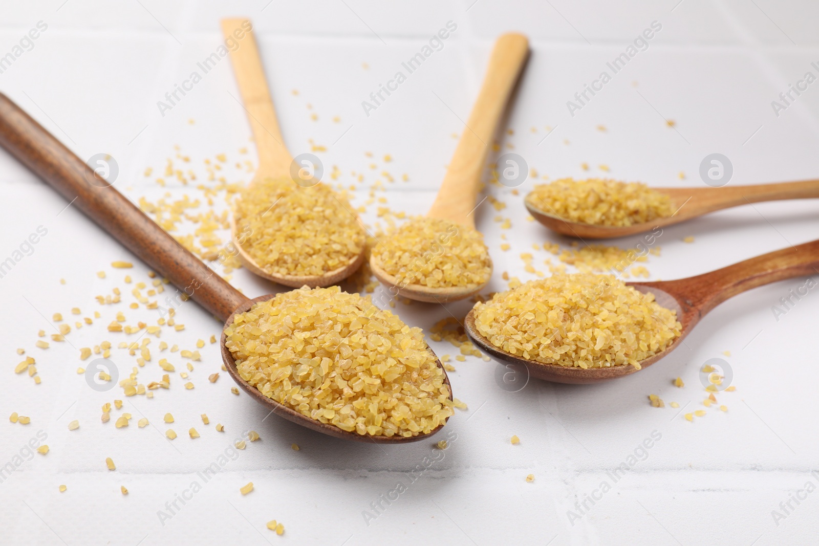 Photo of Spoons with raw bulgur on white tiled table, closeup