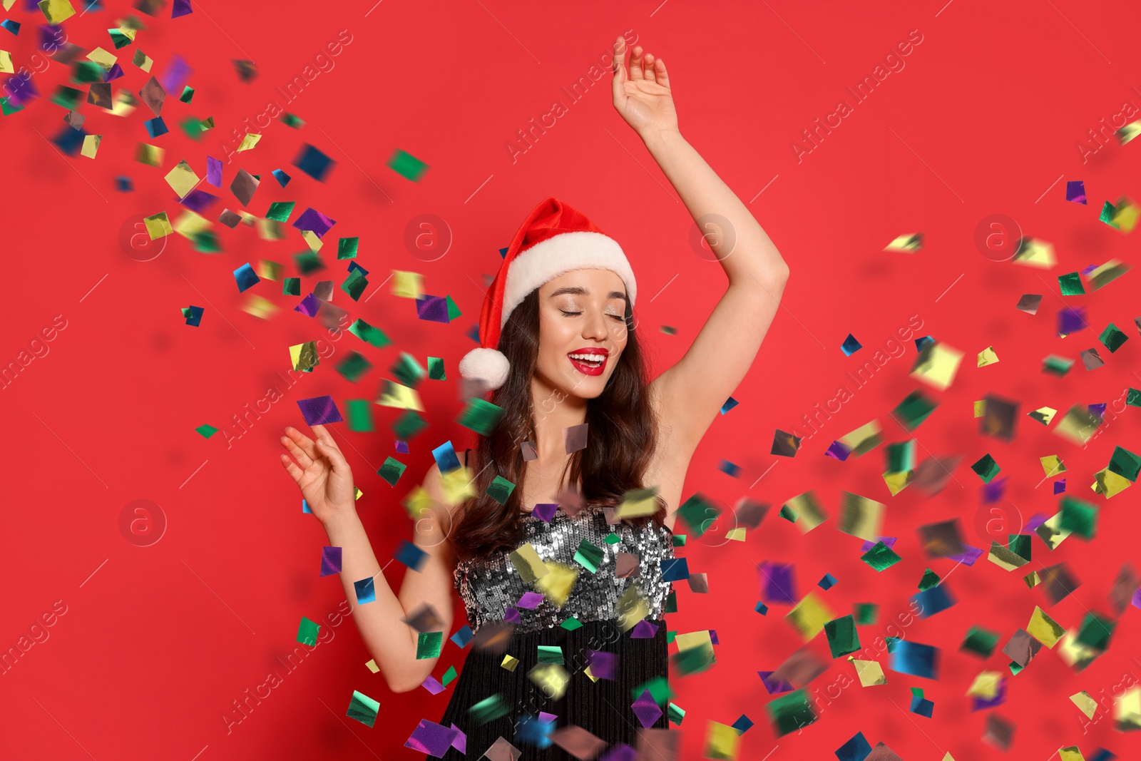 Image of Happy woman in Santa hat with Christmas gift on red background. Colorful confetti flying near her