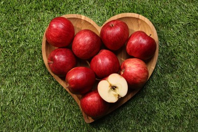 Photo of Wooden plate in shape of heart with red apples on green grass, top view