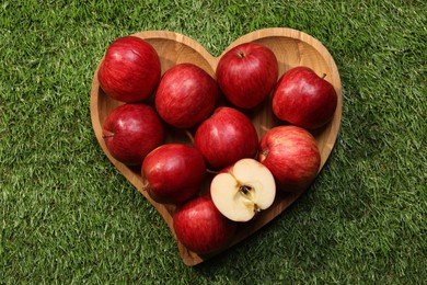 Wooden plate in shape of heart with red apples on green grass, top view