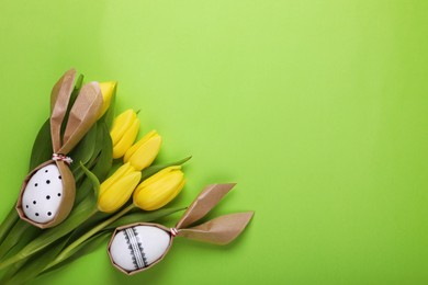 Easter bunnies made of craft paper and eggs among beautiful tulips on light green background, flat lay. Space for text