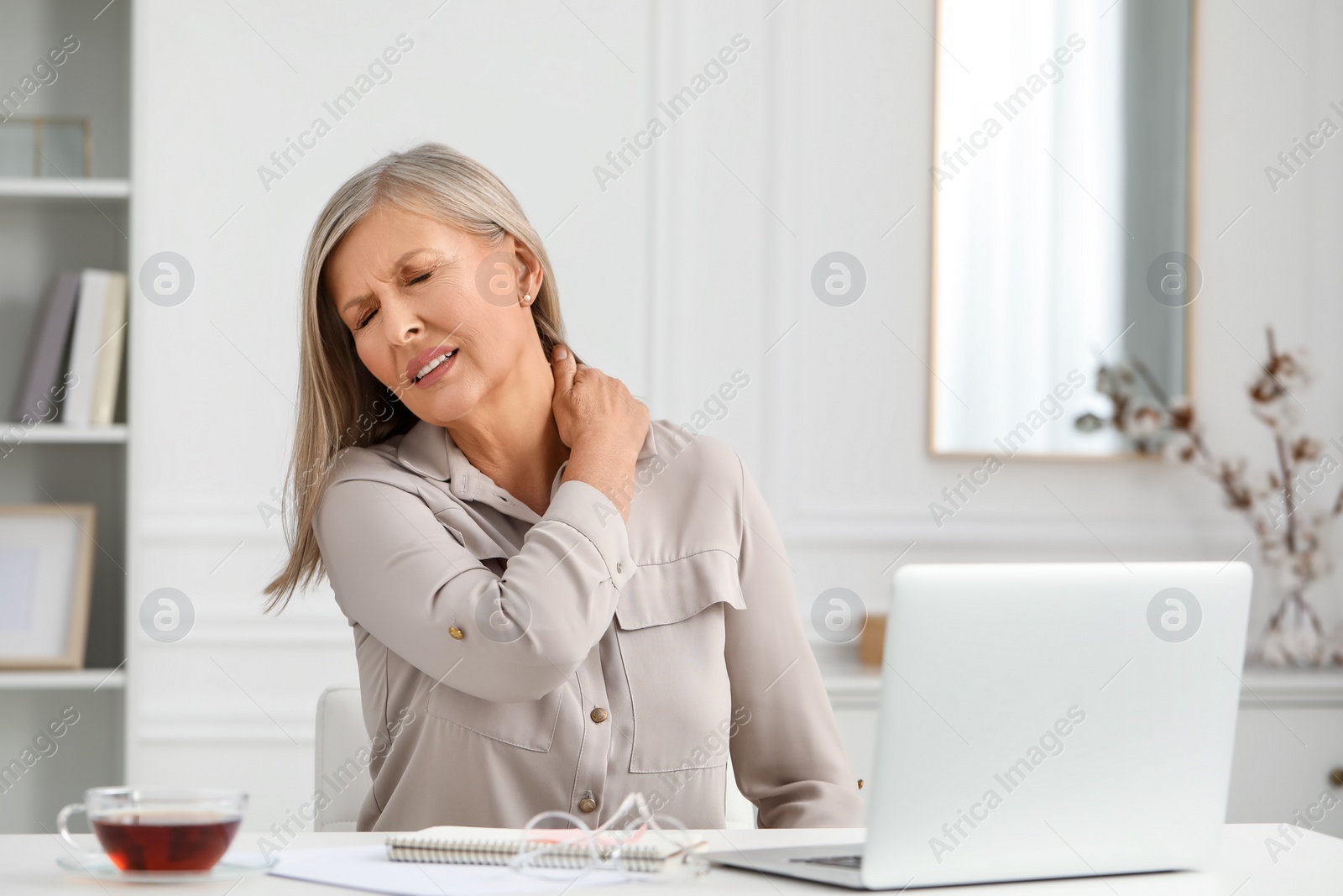 Photo of Woman suffering from neck pain at workplace in room