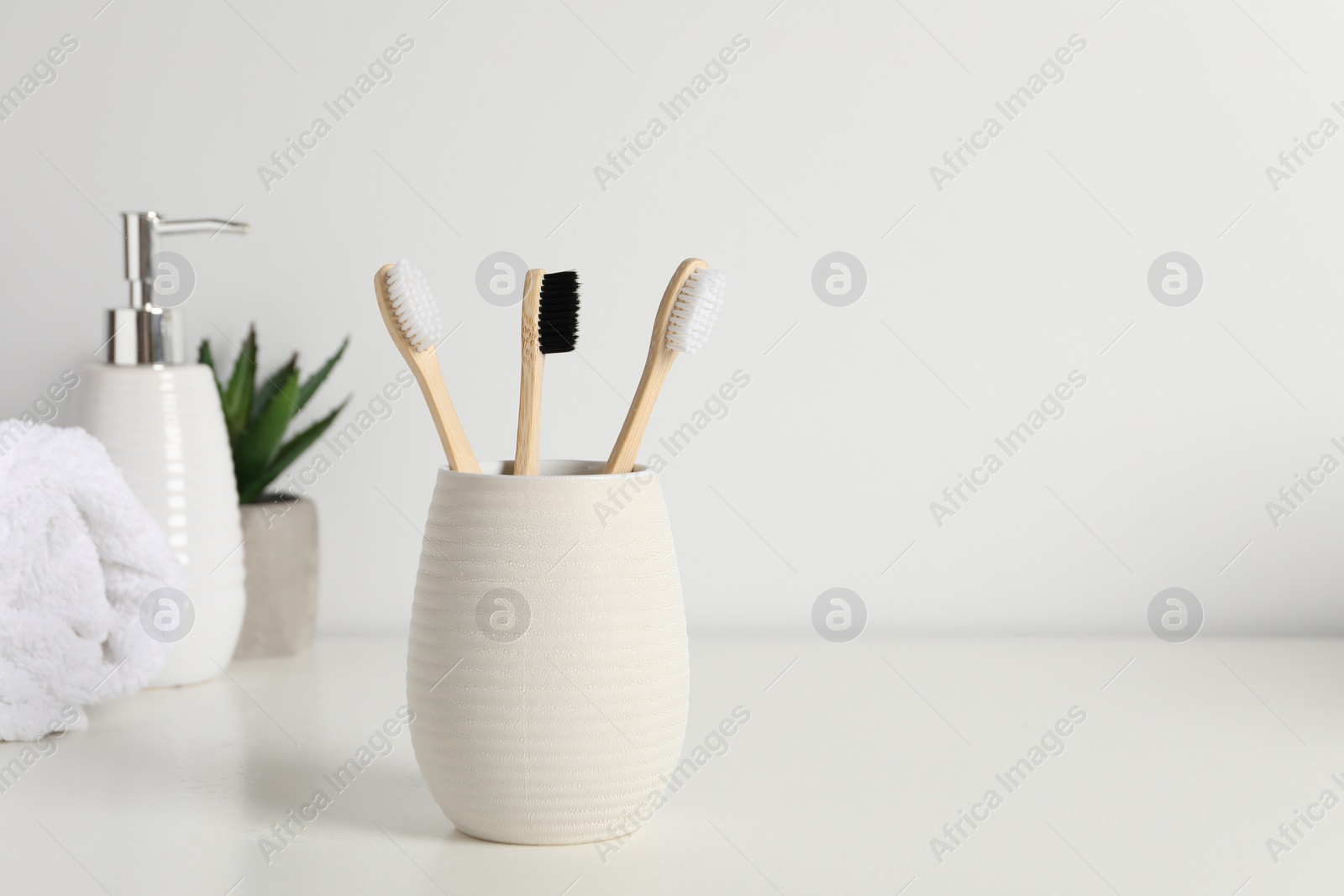 Photo of Bamboo toothbrushes in holder, potted plant, towel and cosmetic product on white countertop, space for text